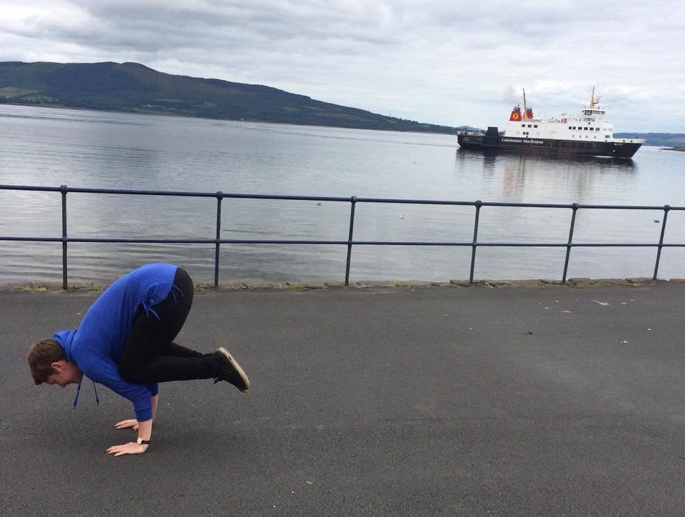 Jennifer Ellis in crow pose on the Ayrshire coast, a Caledonian MacBrayne ferry passing behind
